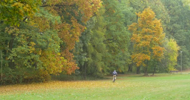 Человек в теплой шляпе, шляпа Ушанки с меховыми ушами Человек с рюкзаком едет на велосипеде по лугу в лес осенью сошел с велосипедной прогулки — стоковое видео