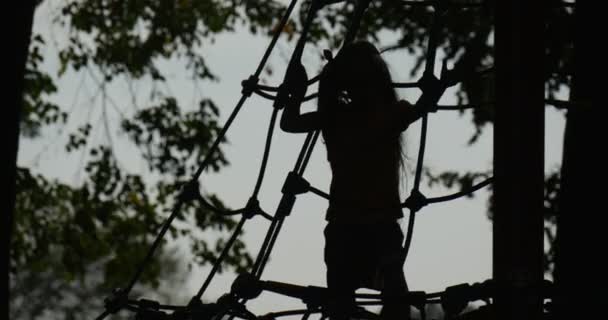 Meisje Close Up is klimmen naar de touw trap meisje van silhouet weinig meisje met lange eerlijke haren in T-Shirt Orange speelt op Speeltuin boomstammen — Stockvideo