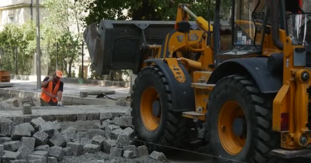 Conductor en la excavadora amarilla descarga el polvo de granito a los bloques Reparación de carreteras City Street Workers in Orange Workwear are Paving the Road Sunny Day — Vídeo de stock