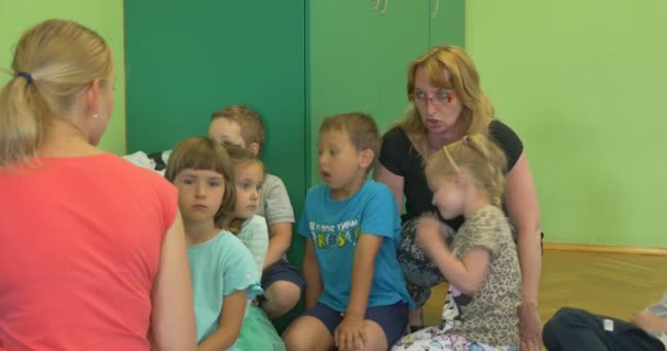 Группа счастливых детей с двумя женщинами-педагогами сидит и играет в классе детского сада Учитель трогает девочку нежно — стоковое видео