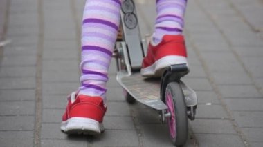 Kız bir tekme Scooter kıza etek ile sarışın saç içinde bir tekme Scooter sokak kızın geri bacaklar kırmızı spor ayakkabılar insanlar kadın anne yavaş hareket içinde Ridding olduğunu