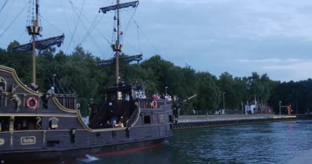 Black Sailboat With People On The Board Fast Swims Into The Harbor Back View Pessoas caminham no cais Água calma Céu Nublado Noite de Verão Leba Poland — Vídeo de Stock