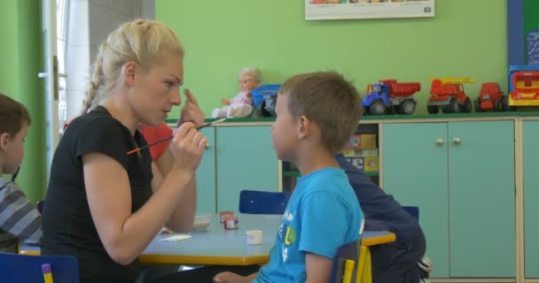 Молодая учительница-блондинка рисует лицо мальчика с раскрашенными лицами смотрит на учителя, играющего в классе детского сада — стоковое видео