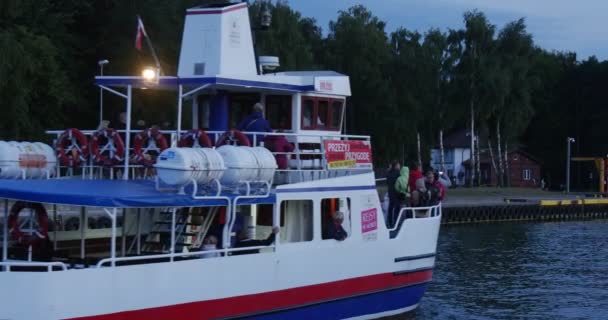 Röd-blå motorbåt med människor på fören simmar in i hamnen nära bakifrån folk går på kajen molnig himmel Twilight sommar kväll Leba Polen — Stockvideo