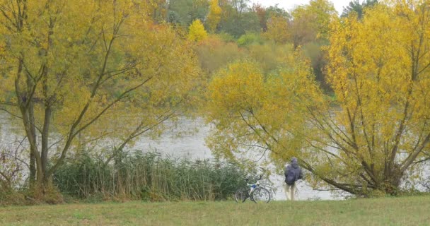 L'homme à Ushanka chapeau et veste marche par la rive du lac vers l'eau En regardant le vélo de l'eau est à gauche sur les arbres verts jaunes herbe 