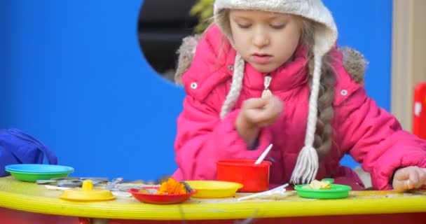 カラフルなプラスチックのおもちゃ料理赤黄色のプラスチックのおもちゃキッチン テーブル子供の遊び場、屋外で遊ぶピンクの暖かいジャケットと白い帽子の少女 — ストック動画