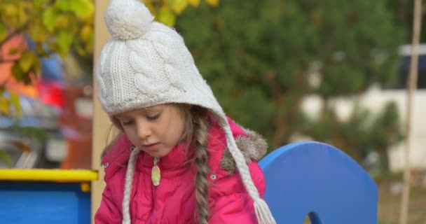 Petite fille joue sur le terrain de jeu dans la cour fille en veste rose et chapeau blanc d'autres enfants courent le dernier jour d'automne en plein air — Video