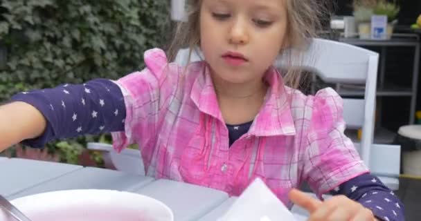 Niña con camisa rosa a cuadros se sienta en la mesa toma servilleta de papel y se limpia la boca — Vídeo de stock