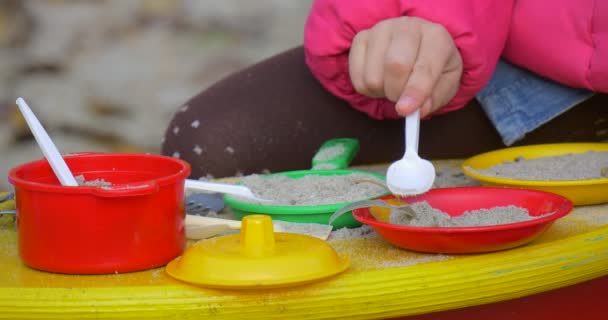 Mãos de menina com colher PLastic Vista Fechar Menina joga com coloridos pratos de brinquedo no quintal ela coloca areia no dia de outono de utensílios de cozinha de brinquedo ao ar livre — Vídeo de Stock