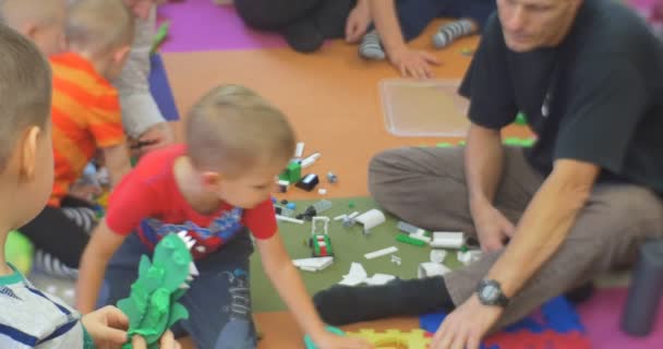 Petits enfants avec leur professeur mâle sont assis plancher rampant regardant et jouant un constructeur de jouets en plastique à la bibliothèque centrale enseignant ouvert un livre — Video