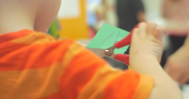 Kisfiú vágás olló egy zöld darab papírt, hogy elmosódott papír Toy-emberek a háttérben központi könyvtár Workshop játékok létrehozása — Stock videók
