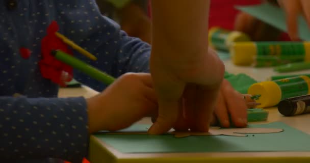 Mała dziewczynka w niebieskiej koszulce jest malowanie na zielonym kawałku papieru przez zielony marker nauczyciel pomaga ręce bliska Centralna Biblioteka warsztaty tworzenia zabawek — Wideo stockowe