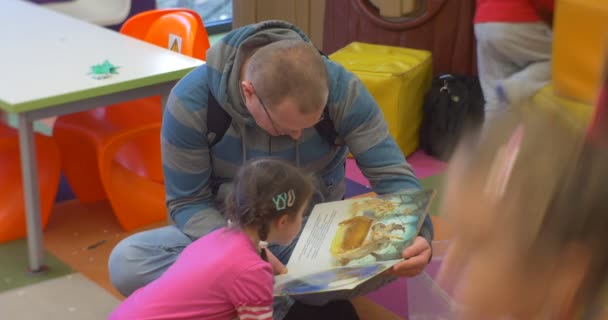 Öğretmen veya Baba çocuksu kitap erkek ile bir Zemin Yapraklama Oturan Küçük Kız Oyuncak Oluşturma Merkez Kütüphane Atölyesi'nde Sınıf Geldi — Stok video