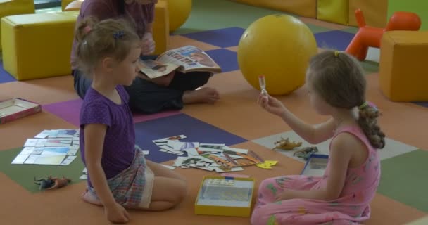 Dos chicas rubias están sentadas tumbadas en un piso jugando al profesor está leyendo libro infantil Los niños están jugando en el aula en la Biblioteca Central en Opole Polonia — Vídeos de Stock