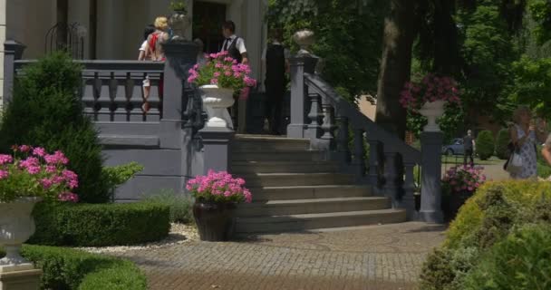 夫妻新娘和新郎走楼下行走轮妇女穿着白色裙子服饰新娘巡游，绿色公园老式建筑中的人 — 图库视频影像