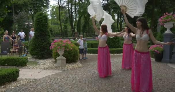 Tres mujeres en trajes de danza del vientre Sujetador blanco Faldas rosadas están bailando con abanicos blancos Balanceándose por los fans La gente está mirando sentado en Green Park Escaleras — Vídeo de stock