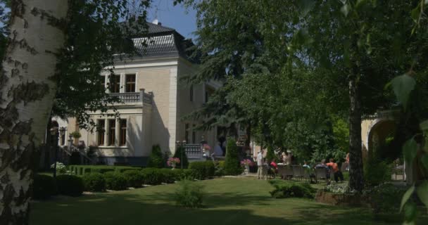 Menschen gehen in Richtung Vintage House White Building im Park Menschen gehen, um eine Bräute Parade grüne üppige Bäume und dekorative Sträucher sonnigen beobachten — Stockvideo