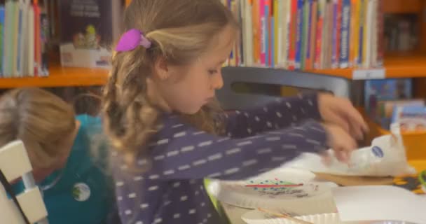 Kleines Mädchen sucht zwischen Papier etwas Malendes am Tisch Mädchen mit zwei Zöpfen Pädagogin Frau spricht mit jemandem Bücherregale auf dem Hintergrund — Stockvideo