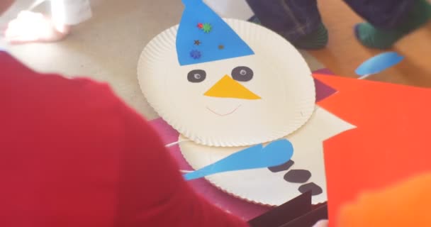 Deux enfants sont assis avec leurs bonhommes de neige plancher de salle de classe finissant leurs appliques faisant un bonhomme de neige d'une assiettes jetables et du papier Applique du Nouvel An — Video