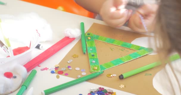 Le mani dei bambini decorano un triangolo con perline e paillettes Le mani da vicino I bambini sono seduti a tavola I marcatori colorati sono sul tavolo — Video Stock