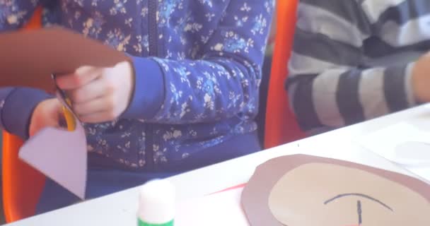 Παιδί κόβει ένα καφετί φύλλο χαρτί από ψαλίδι για δύο παιδιά κάθονται στο τραπέζι χαρτί κοπεί απλικέ είναι σε ένα πίνακα εκπαιδευτικός έρχεται σε βοήθεια — Αρχείο Βίντεο