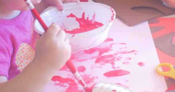 Niña pequeña está pintando con pintura rosa pone un pincel en un tazón con pintura y pone una mancha en una hoja blanca de papel corte tijeras de papel en una mesa — Vídeo de stock