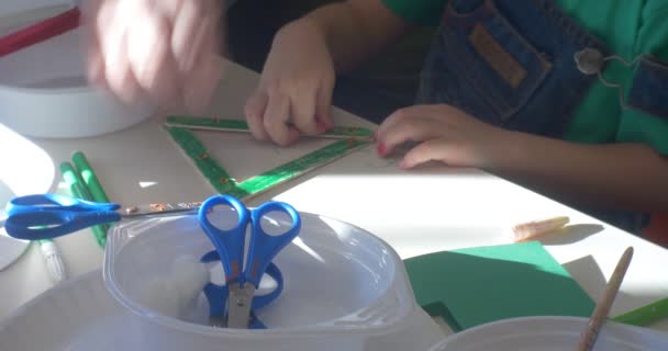 Маленький мальчик-воспитатель украшает треугольник, окрашенный в зеленый цвет, украшенный бусами, руки людей сближают Мальчик и женщина режут ножницы бумаги, зажженные солнцем — стоковое видео
