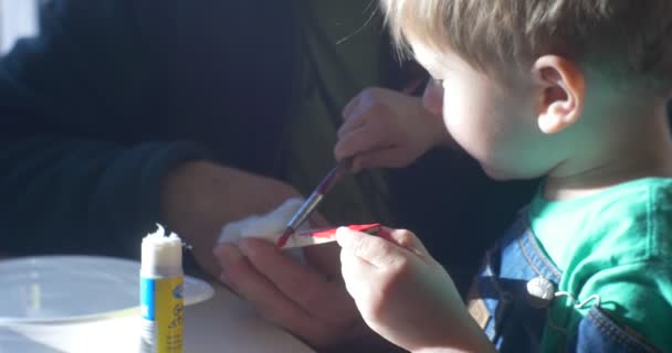 Kleine jongen in groen T-Shirt is schilderen door het penseel Kid's gezicht verlicht door de zonnestralen Kid en opvoeder maken een New Year's Winter Appliques in een klaslokaal — Stockvideo