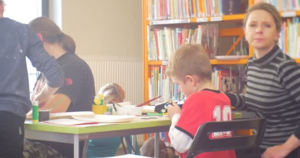 Junge Kind sitzt am Tisch mit einem Erzieher oder einer Mutter, die eine Winterapplikation für das neue Jahr schneidet ein Papier in einem Klassenzimmer Bücherregale — Stockvideo