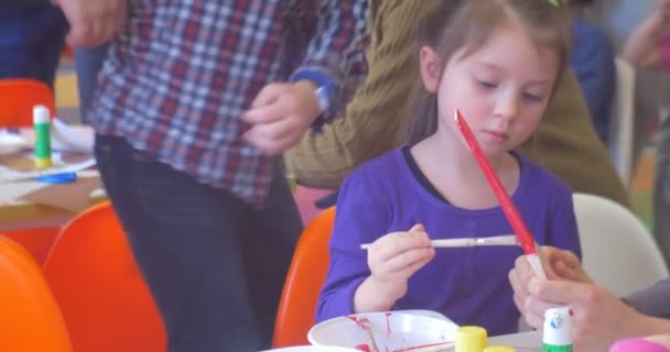Little Girl is Painting a Wooden Spoon Attentively with Red Color By Brush Educator is Holding a Spoon Helps the Girl Kid is Making a New Year 's Gifts — Stok Video
