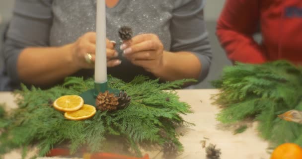 Eine erwachsene Frau bereitet einen Tannenzapfen zu und fügt ihn behutsam zur weihnachtlichen Komposition von Adventskranzzweigen am Tisch hinzu. — Stockvideo