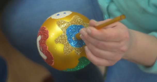 Weibliche Hände Frau in blau hält goldenes Spielzeug Neujahrsbaumkugel steht auf und geht weg Ball ist mit verschiedenen Farben glitzert bemalt — Stockvideo