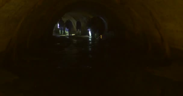 Los turistas exploradores están caminando lejos en una cueva con lámparas Semicircular Tunnel Lit by Lamps Small Water Stream is Under People 's Feet Underground River — Vídeos de Stock