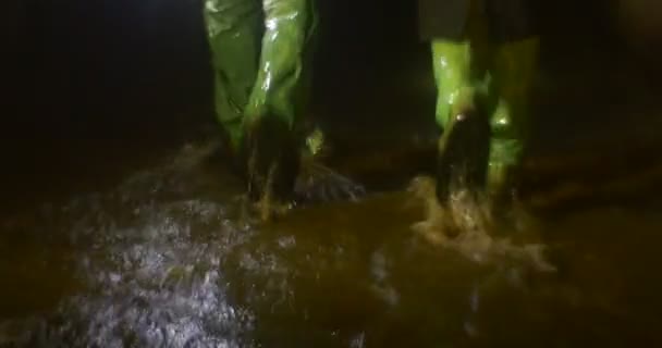 Casal está segurando as mãos Homem com mochila Grupo de turistas está caminhando para a frente por uma caverna escura em botas verdes por uma corrente de água Rio subterrâneo — Vídeo de Stock