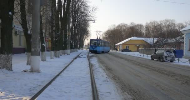 Blaue Straßenbahn nähert sich Bahnhof Fußgänger gehen durch eine Straße alte Straßenbahn bewegt sich durch eine Eisenbahn Winter Gebäude kahle Äste Bäume — Stockvideo