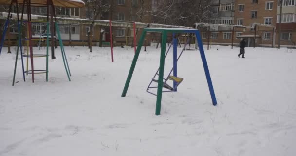 Человек идет через двор качели песочница игровая площадка оборудование покрыто снегом детской площадки в зимних жилых домах много снега — стоковое видео