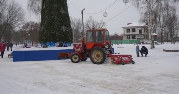 Trator se move ao longo de uma árvore de ano novo no parque Konotop Red Tractor está limpando um beco do parque remove a neve pessoas andando ramos nus árvores — Vídeo de Stock