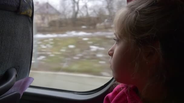 Dziewczyna jest obserwowanie wsi domy gołe gałęzie drzew blondynka dziewczynka jest patrząc przez okno w autobus z jej twarzy spokojnej krajobraz jest zmiana — Wideo stockowe