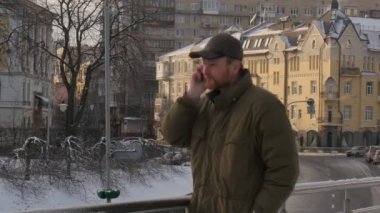 Adam ile sakal Talking tarafından telefon ve gülümseyen duran bir Bridge Winter Steam Comes Off a Mouth Vintage Brown-Blocked Bina Cityscape arabalar yolda olduğunu