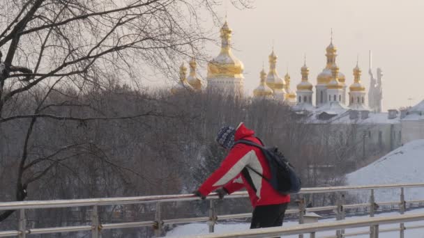 El hombre de rojo corre y patina en un pedazo de puente peatonal de hielo en Wintery Park El hombre se detiene inclinado a los rieles y las torres de espera Kievo-Pecherskaya Lavra — Vídeo de stock