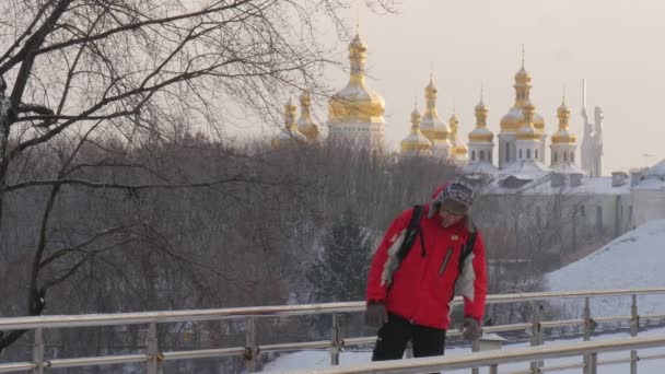 Hombre está de pie se inclinó a un pie de rieles de puente durante un tiempo y ejecutan a Backpacker en wintry Parque oro cúpulas Kievo-Pecherskaya Lavra en un horizonte — Vídeos de Stock