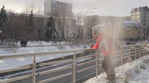 Mann sammelt Schnee von einem Boden wirft Schnee von einer Brücke Mann Backpacker ist auf einer Brücke im Winter moderne und alte Gebäude am ruhmreichen Ort — Stockvideo
