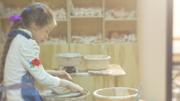 Chica está moldeando un círculo con precisión con espátula trabajando en una rueda de cerámica Niña rubia se esfuerza por hacer un regalo de una arcilla en taller de cerámica — Vídeo de stock