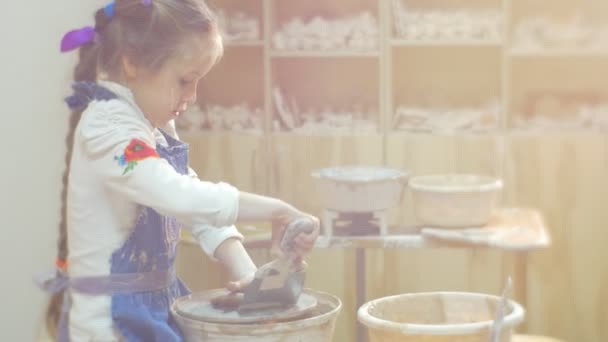 Девушка формирует круг точно с лопаткой, работающей на гончарном круге Маленькая блондинка старается сделать подарок из глины в мастерской керамики — стоковое видео