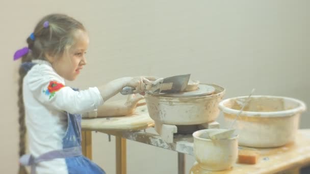 Fille est en train de mouler un cercle sur une roue de poterie tenant une spatule tournant une roue par jambe Petite fille blonde essayant dur de faire un cadeau à partir d'un atelier d'argile — Video