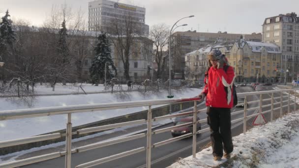 Ο άνθρωπος είναι μιλώντας από κινητό τηλέφωνο αφαιρεί ένα χιόνι από ένα Backpacker άνθρωπος ράγες είναι σε γέφυρα στο χειμώνα χιόνι σύγχρονο και Vintage κτίρια στο δόξα τόπος Κίεβο — Αρχείο Βίντεο