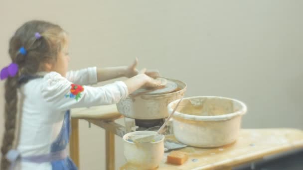 La fille tourne une roue de poterie concentrée par moulage de jambe une petite fille blonde en argile fait des efforts pour faire un cadeau à partir d'une argile dans un atelier de poterie — Video