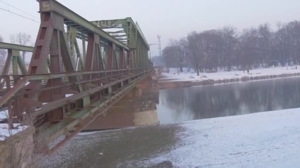 Leere Brücke alte Eisenbahnbrücke durch einen Fluss Metallkonstruktionen Häuser am gegenüberliegenden Ufer glattes Wetter winterliche Landschaft Schnee auf dem Boden — Stockvideo