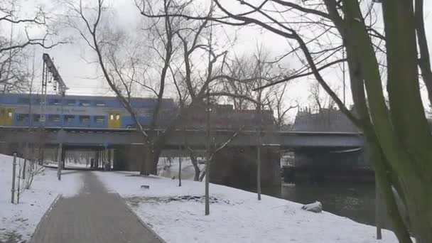 Vonat túrák át a híd lassított gyalogos járda alatt egy híd révén egy kis folyó lombtalan fák Park, a folyó a téli Kijevben — Stock videók