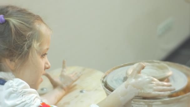 Niño está girando una rueda de cerámica haciendo esfuerzos moldeando un florero Niña rubia está trabajando en una rueda de cerámica concentradamente haciendo un regalo en el taller — Vídeo de stock